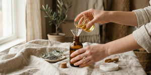 Person pouring liquid from a glass bottle into an amber glass bottle using a funnel on a textured surface.
