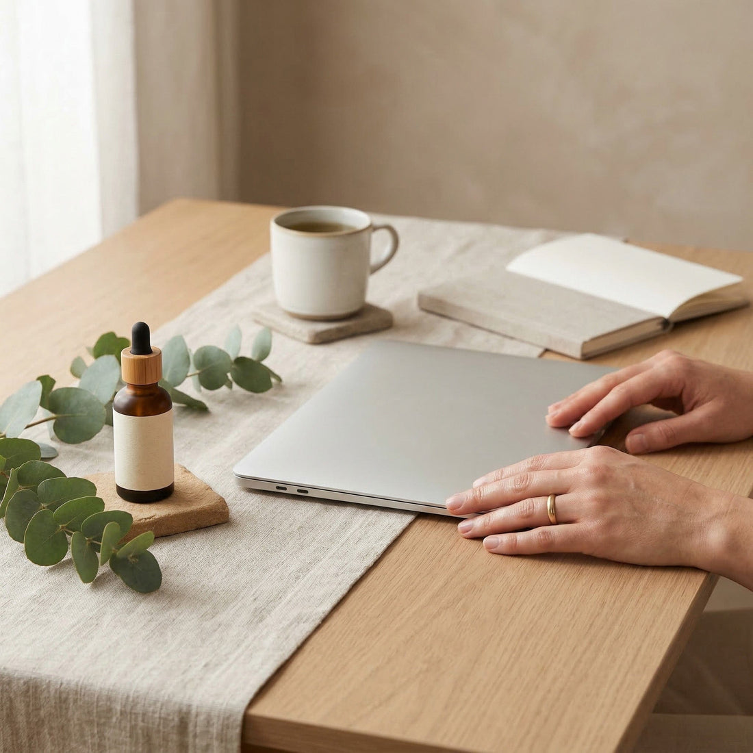 Person interacting with a laptop on a wooden table with a cup, book, and plant.
