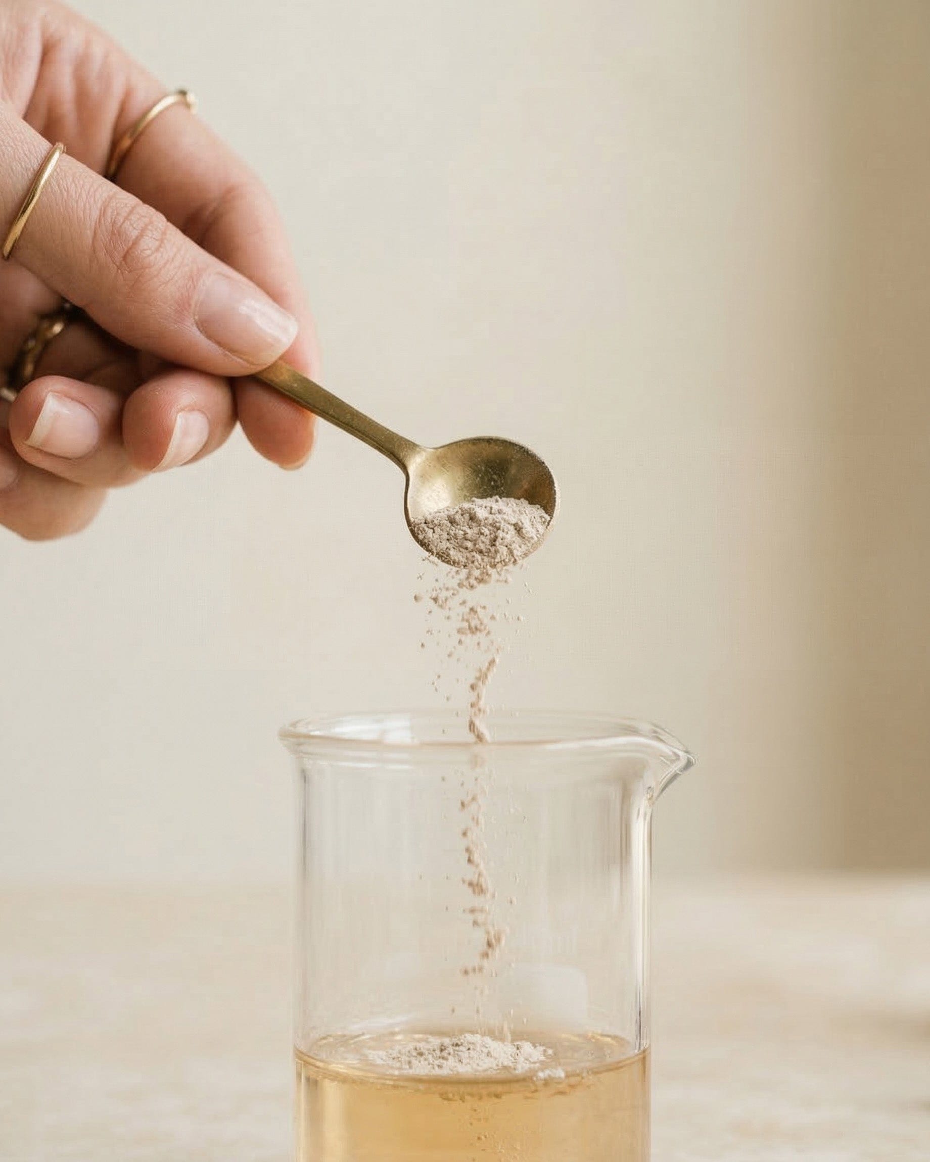 Hand pouring a spoonful of a light brown powder into a glass container with a neutral background.